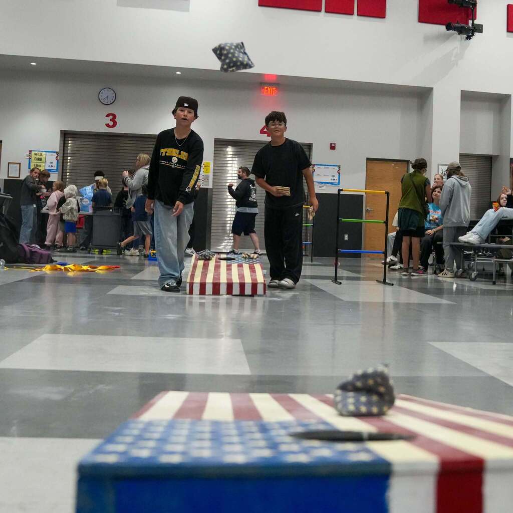 Image 10: Two students playing cornhole inside a school cafeteria or commons area, with a beanbag mid-air as it flies toward an American flag-themed board.