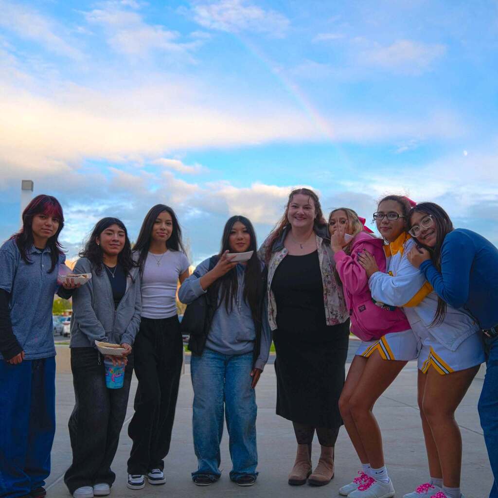 Image 1: A group of high school students and a staff member smiling and posing together outdoors under a light sky with a faint rainbow overhead. Two students on the right are wearing cheerleader uniforms and hugging, while others hold plates of food.