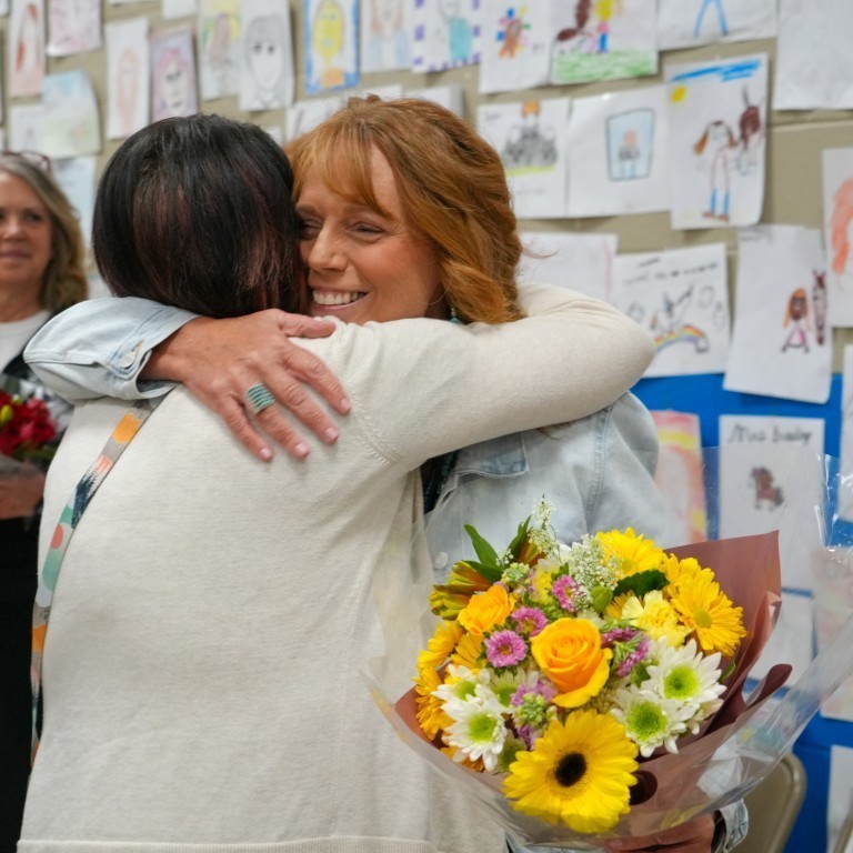 A close-up of Kari Breeding smiling warmly as she hugs a community member while holding a bright bouquet of yellow flowers.