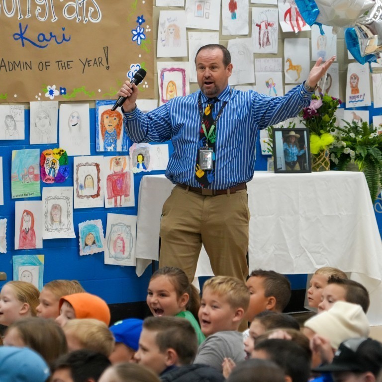 A Weber School District administrator speaking enthusiastically into a microphone in front of a wall covered in student-drawn portraits of Principal Breeding.