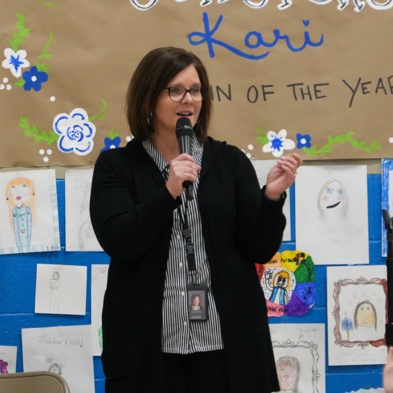 A school staff member speaking at a podium, celebrating Principal Breeding’s legacy in front of a "Congrats Kari" banner.