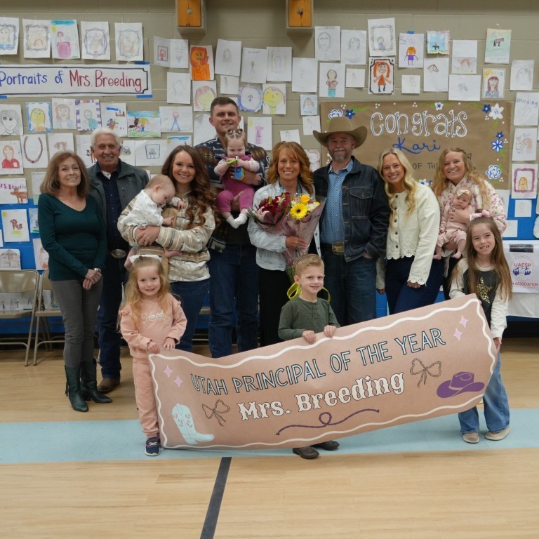 Principal Kari Breeding posing with her family, including children and grandchildren, in front of a student-created "Portraits of Mrs. Breeding" wall.
