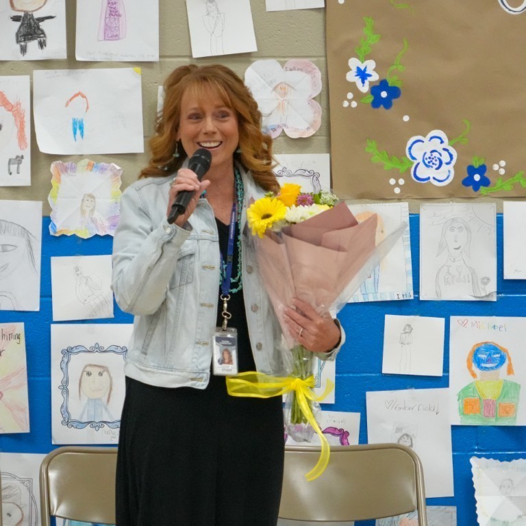Principal Kari Breeding speaking to her students and staff at Hooper Elementary, holding a microphone and a bouquet of flowers.