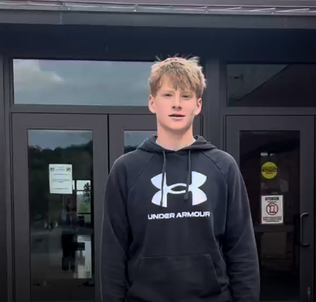 A middle school boy in a black hoodie standing in front of a school entrance.