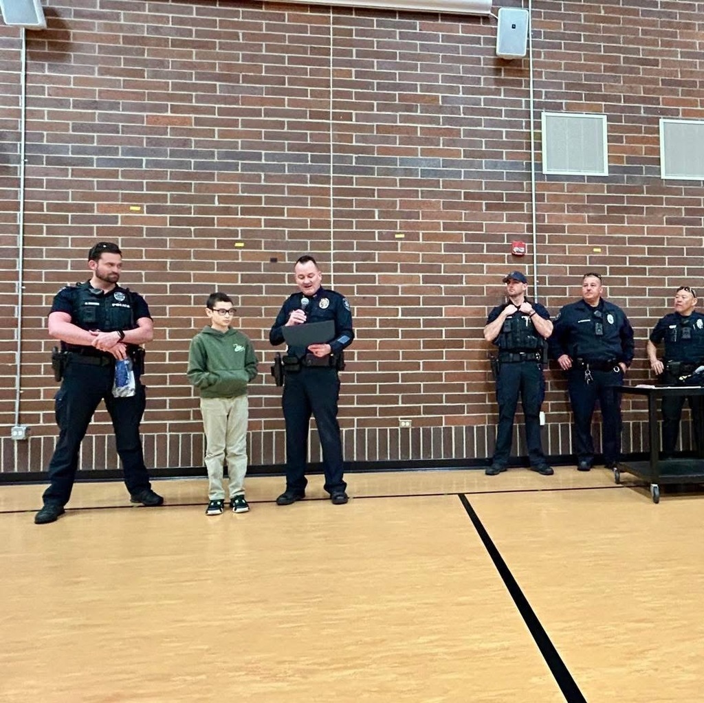 2 police officers with a young boy in front of a brick wall in a gymnasium. One pfficer is speaking into a microphone and awarding the child a certificate