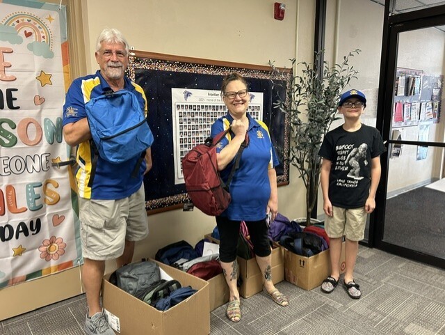 photo of two volunteers from Meridian Optimist, along with a student, holding backpacks and standing in front of several boxes of backpacks
