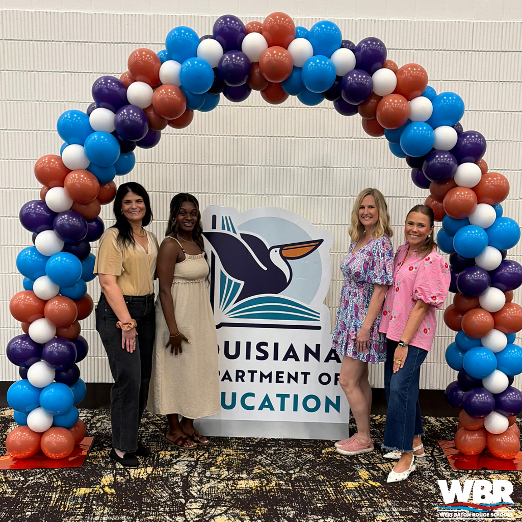 Four women pose for a photo under a colorful balloon arch in front of a sign.