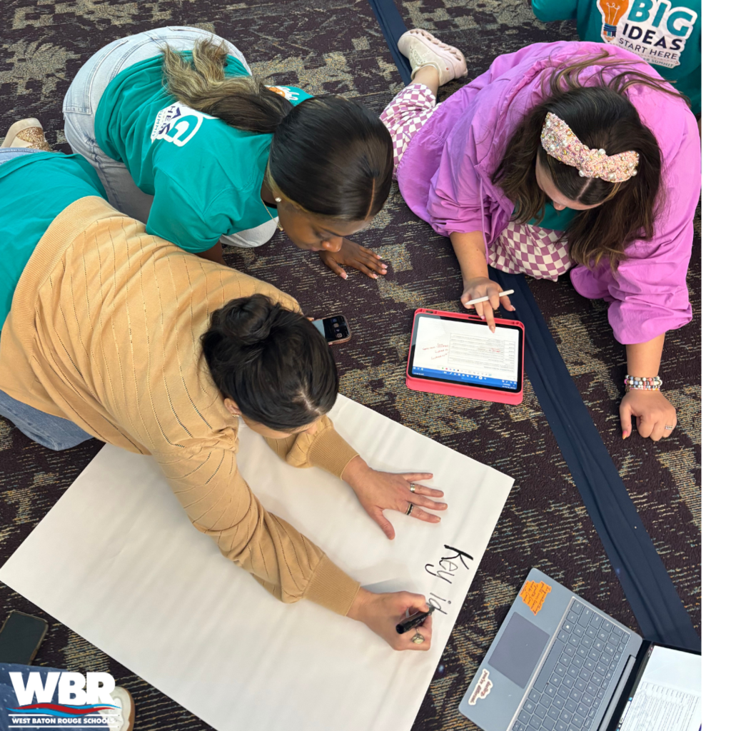 Several individuals work together on a large sheet of paper. One holds a tablet.