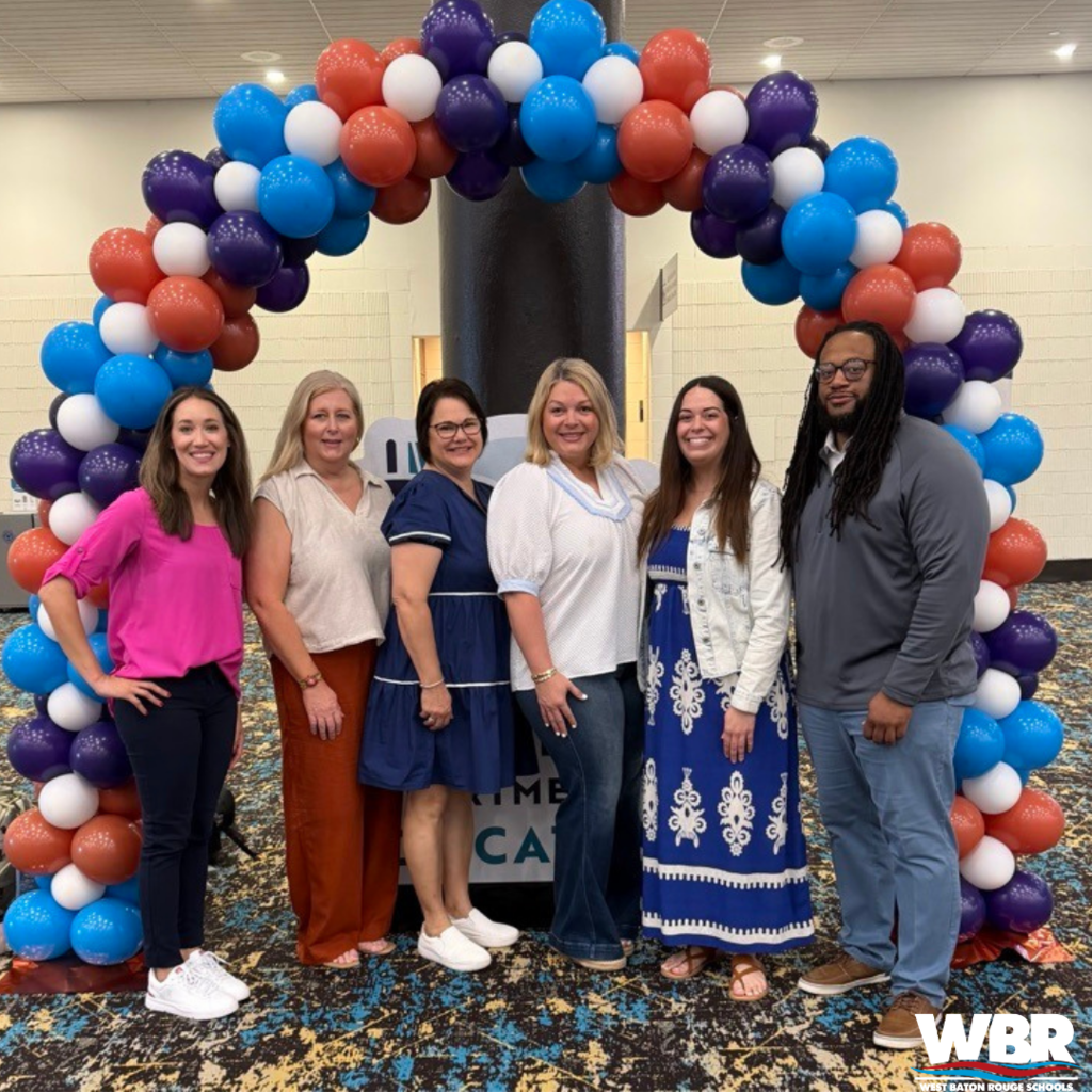 Seven adults pose for a photo beneath a colorful balloon arch in a room with a dark floor.