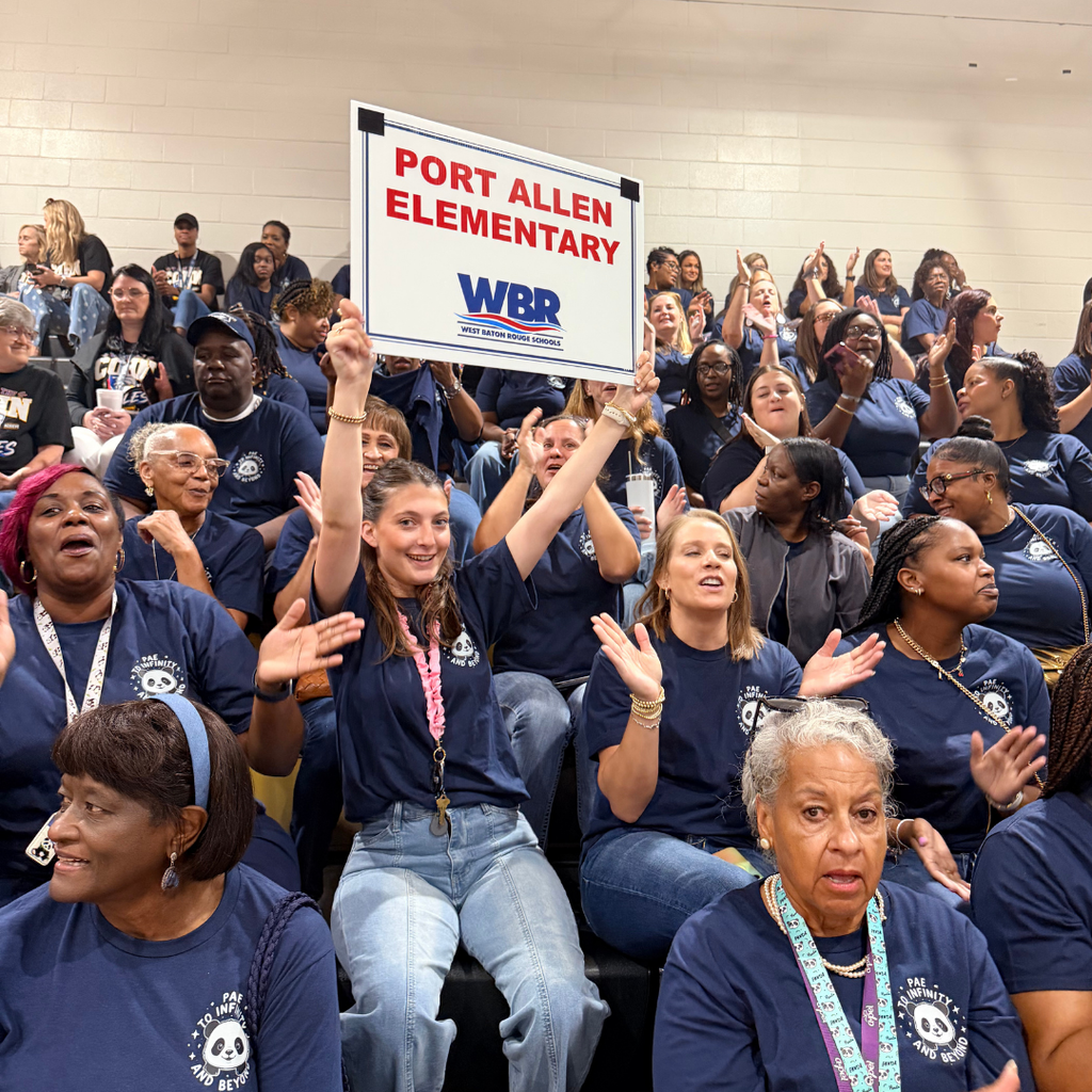 Large crowd of people in blue shirts seated in a room. Person in jeans holds a sign reading "PORT ALLEN ELEMENTARY".