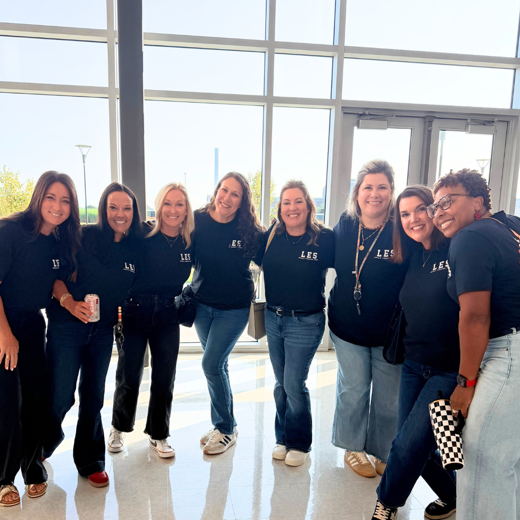 A group of seven women wearing matching shirts stand in a hallway. They pose with smiles.