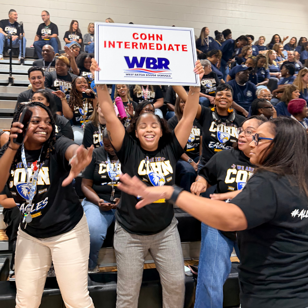 A group of adults and students celebrate together in a school auditorium. A student holds up a sign.