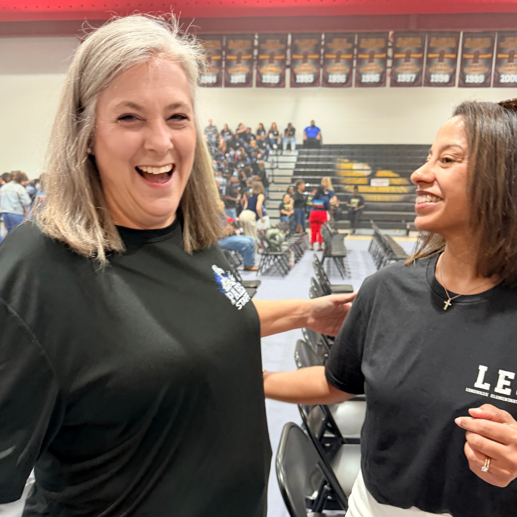 Two women, smiling, stand next to each other in a gym. One wears a shirt with white text.
