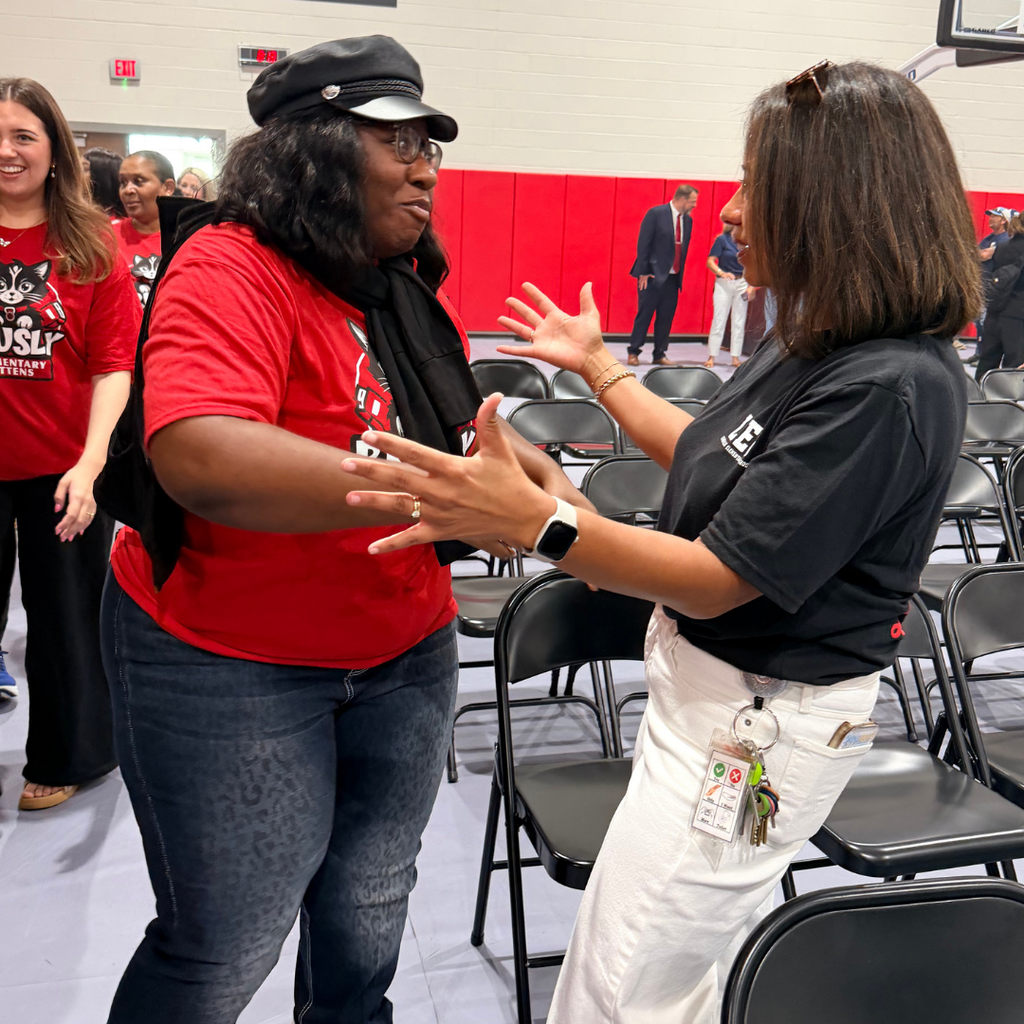Two women converse in a room with rows of chairs. The woman in red gestures with hands.