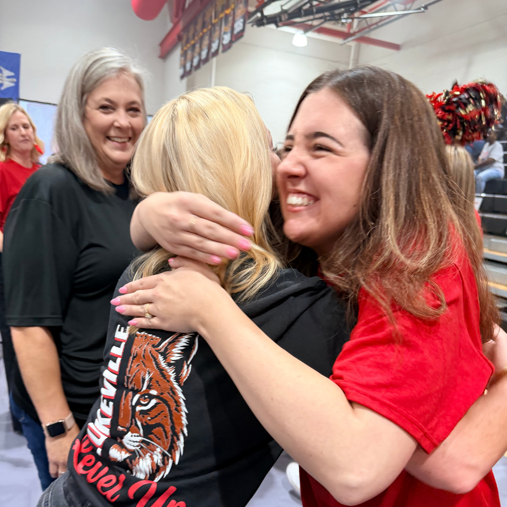 Two women embrace with a smiling woman in red; a woman with blonde hair wears a black sweatshirt.