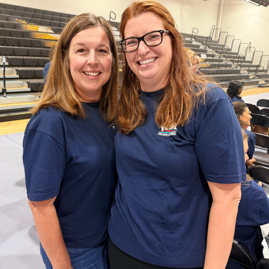 Two women in blue shirts stand close together, smiling and looking at the camera in an indoor auditorium.