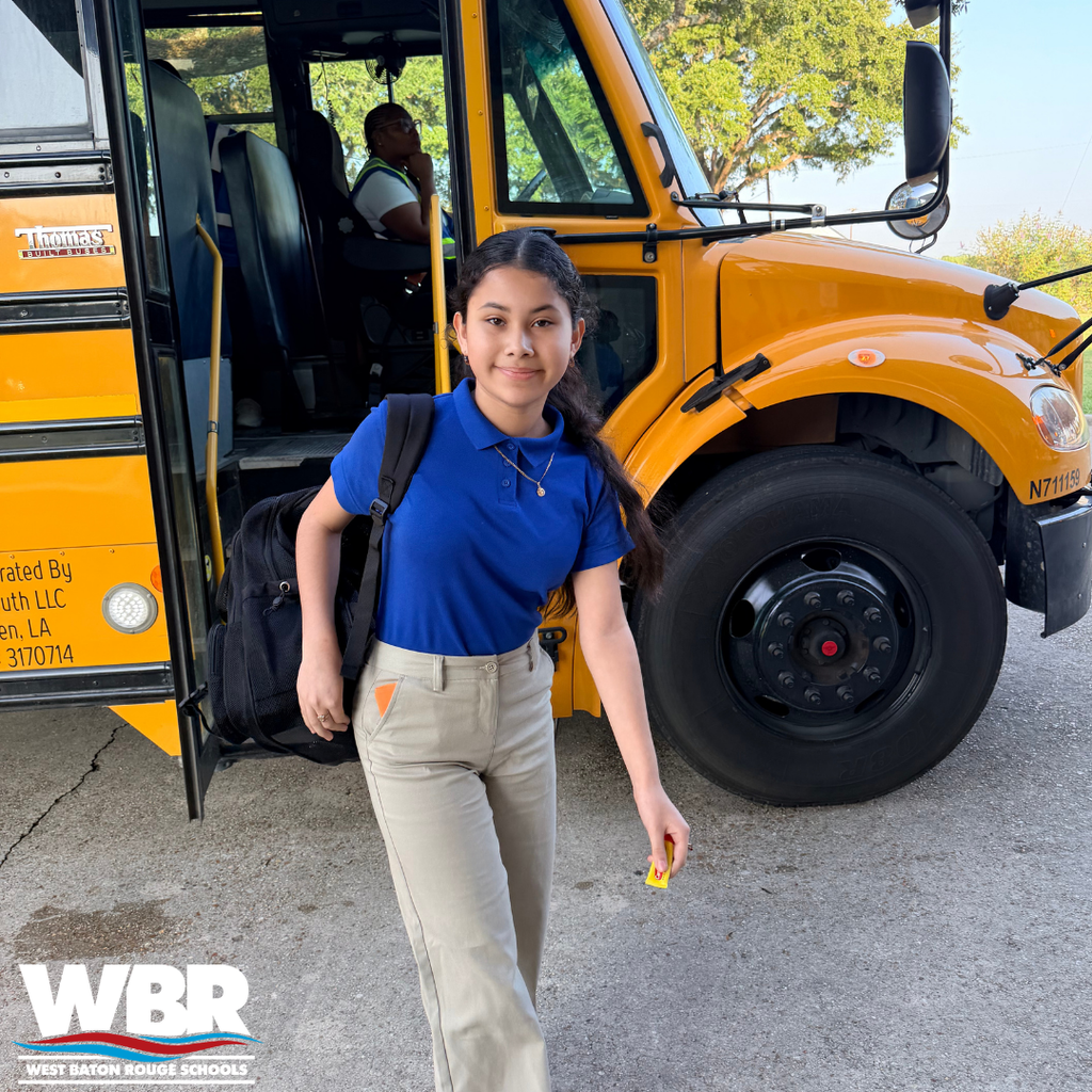 A girl in blue top and beige pants walks by a school bus, holding a yellow object.