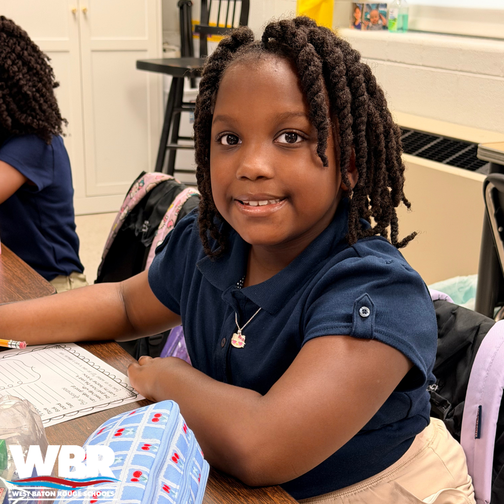 A young girl in a blue shirt sits at a desk in a classroom, smiling, with a backpack behind her.