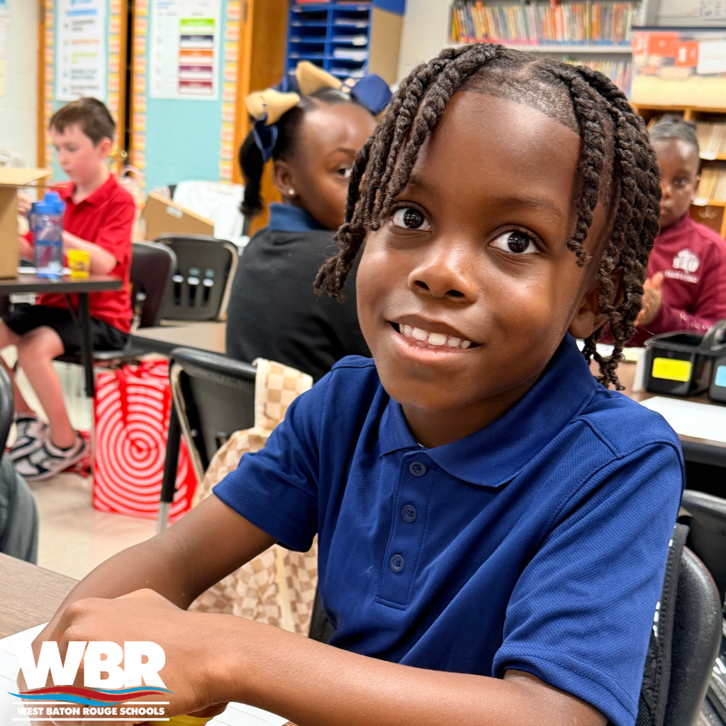 A child with braided hair, in a blue shirt, sitting in a classroom and smiling at the camera.