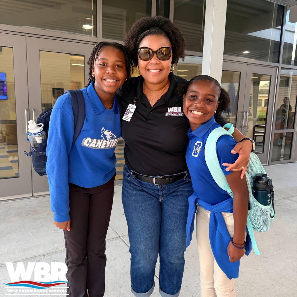 Three individuals stand outside a building. A woman with sunglasses and two girls with backpacks, smiling for a photo.