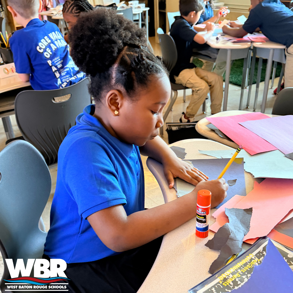 A student in a blue shirt works on an art project with colored paper and a glue stick.