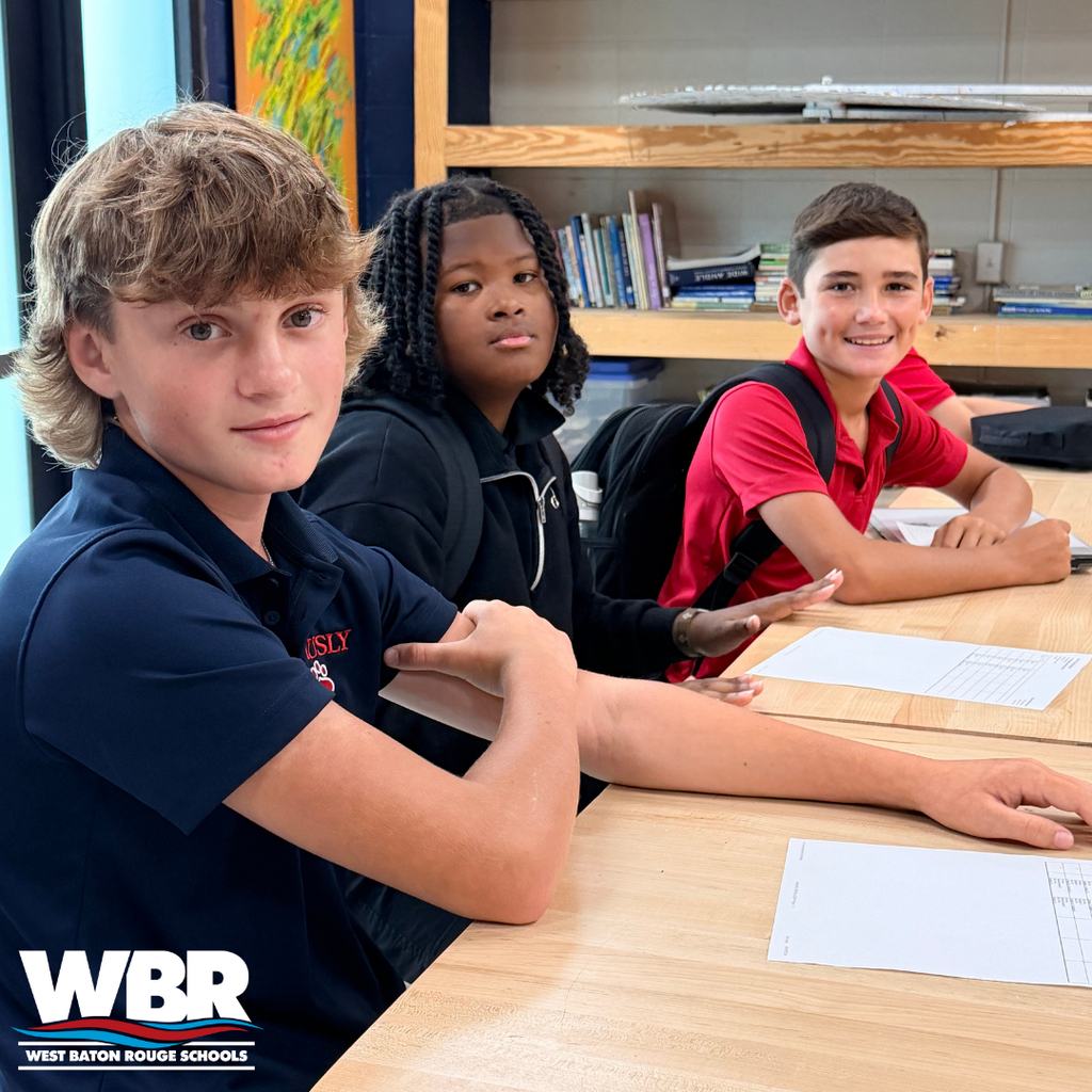 Three students seated at a table in a classroom. The left and middle student are focused on their work, while the right student looks up, smiling.