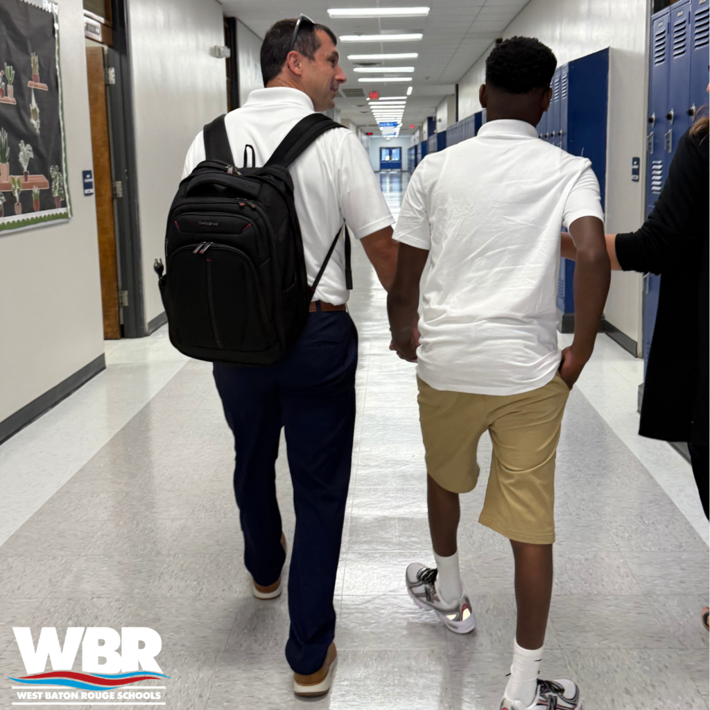 Two men walk in a hallway. One wears a backpack and pants, the other shorts. Both wear white shirts.