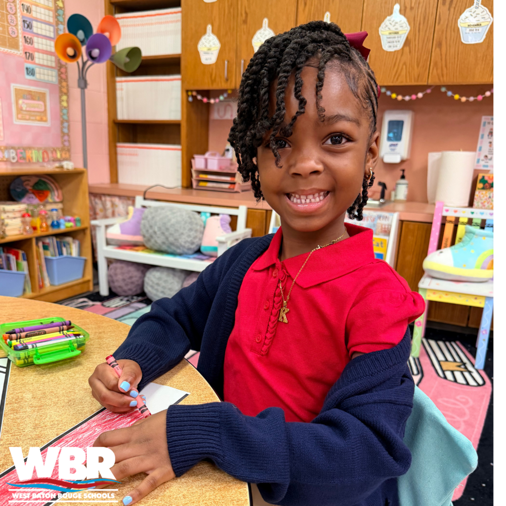 A young girl sits at a desk with a red top and blue sweater, smiling. She holds a marker. Behind, a classroom with shelves, colorful decorations, and a whiteboard.
