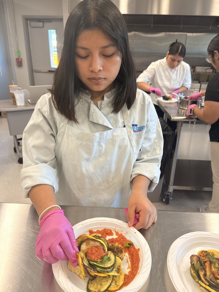 A woman wearing a white chef's coat and pink gloves is cutting vegetables on a plate.