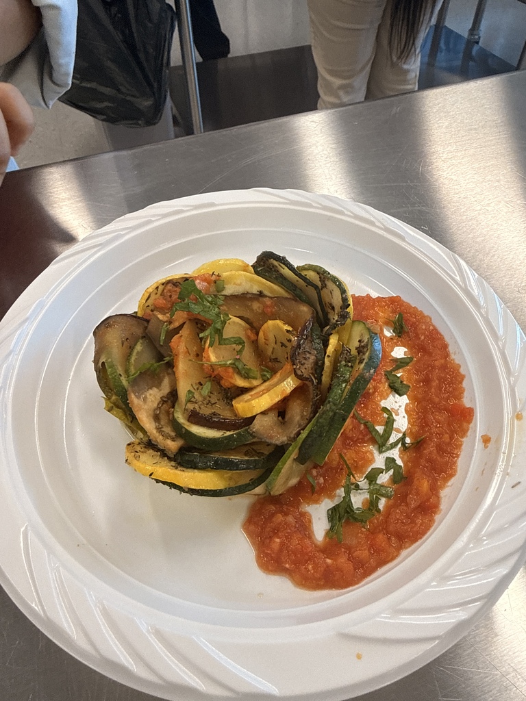 A white plate with a vegetable dish on a metal surface. The dish includes sliced vegetables with a red sauce.