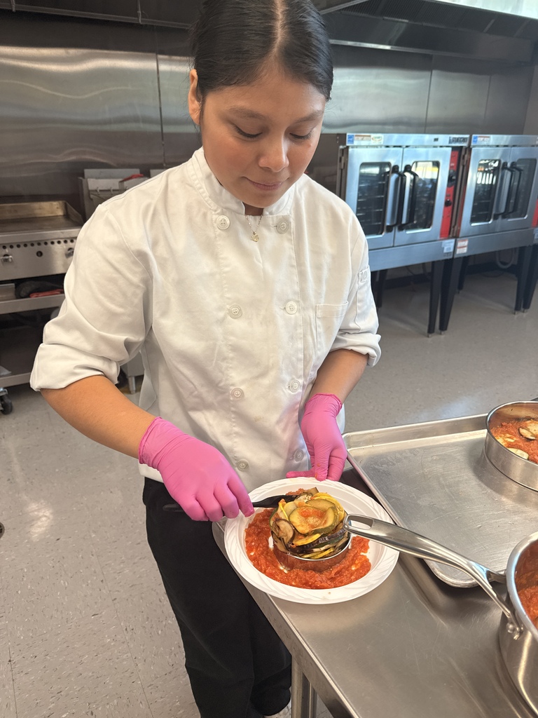 A chef in a kitchen, wearing white uniform and pink gloves, holds a plate with a dish on it.