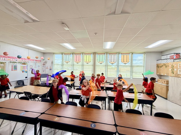 A classroom with children wearing red shirts and holding ribbons, participating in an activity. Tables, chairs, and decorative items are visible.