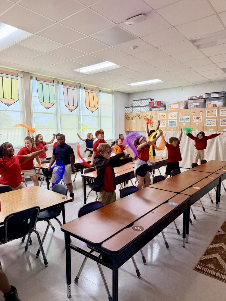 Several children in red shirts and shorts dance in a classroom with desks and tables. Bright sunlight and decorations on the wall.