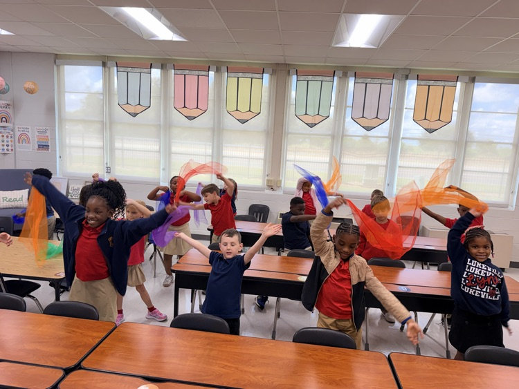 Kids in red and blue shirts dance with colorful fabric in a classroom with large windows.