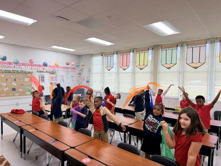A group of children in red shirts stand and play with streamers in a brightly lit classroom.
