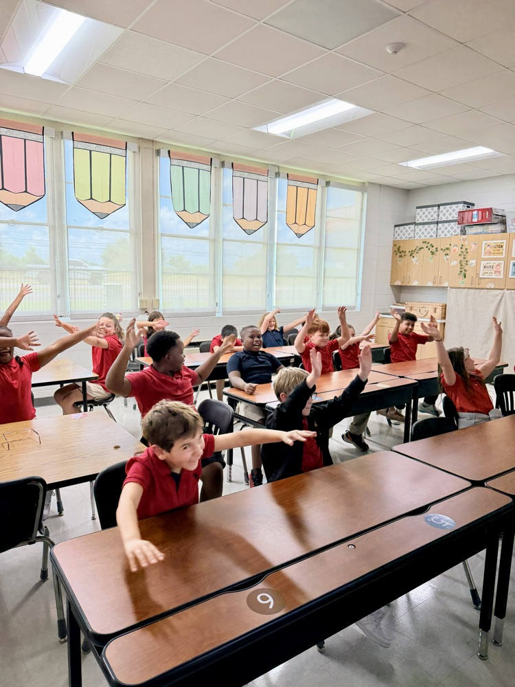 Classroom with children in red shirts seated at desks raising their hands. Bright light from windows.