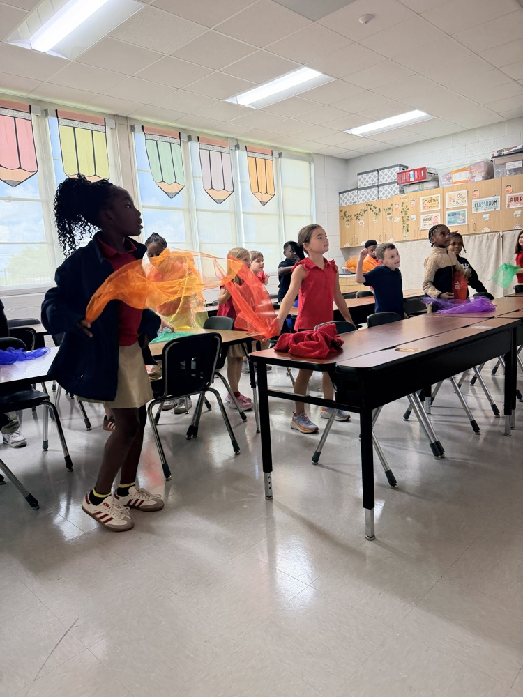 Children in a classroom, one holding fabric while others stand behind desks, with glass windows and fluorescent ceiling lights.