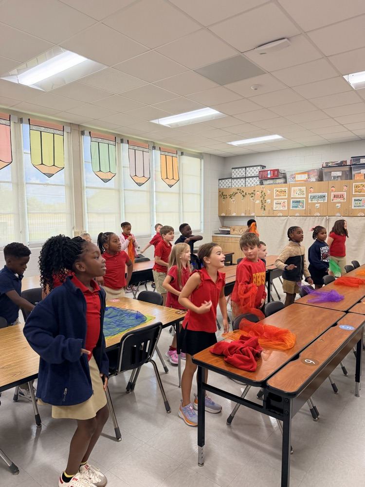 A classroom with children in red shirts and shorts standing and sitting at tables, with colorful curtains on the windows.