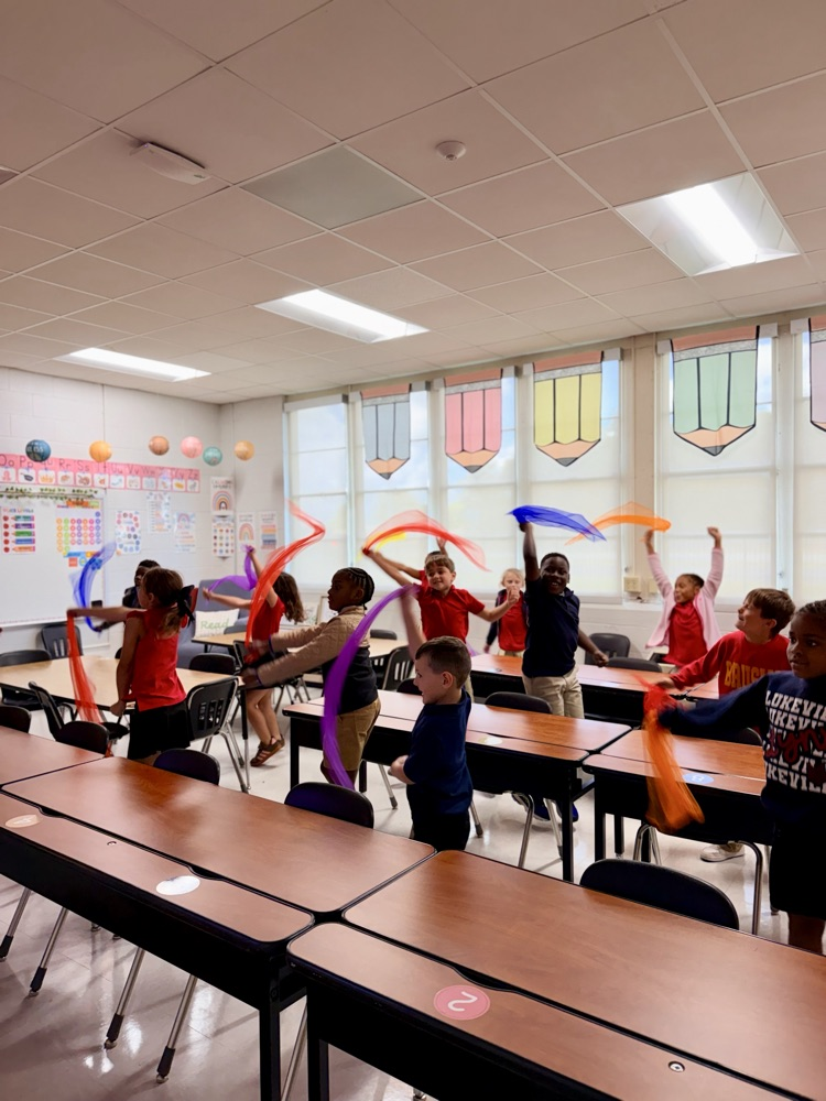 Kids inside a classroom, wearing red shirts, waving around colorful ribbons and plastic hands.