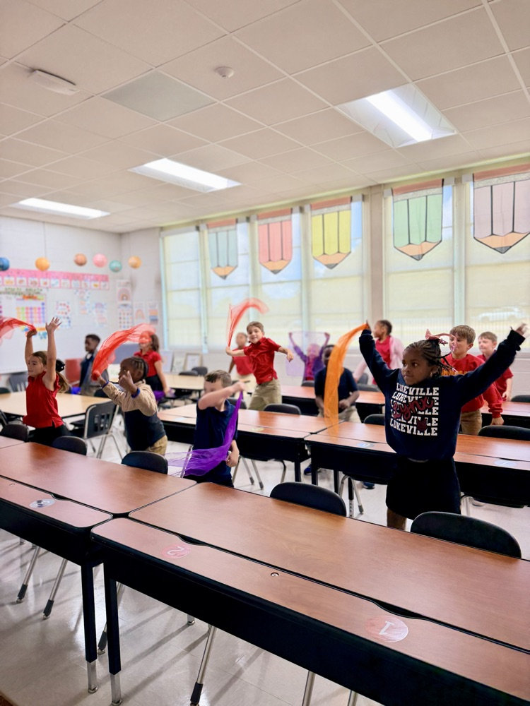 A classroom with desks and chairs, children doing exercises with red ribbons, glass windows with colorful decorations.