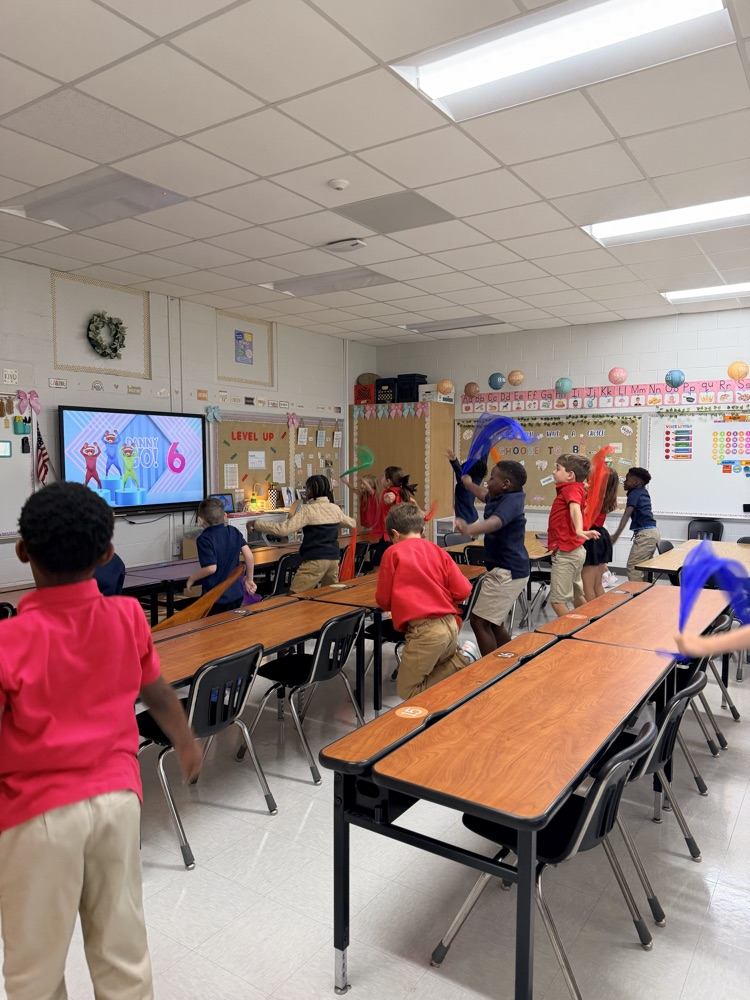 Children in a classroom wave red and blue ribbons. Some sit at tables; a large screen displays a presentation.
