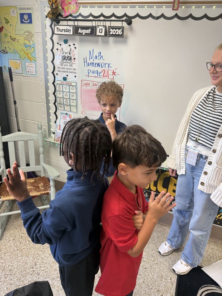 Two boys and an adult woman in a classroom; one boy has his hands up, another holds a sign.