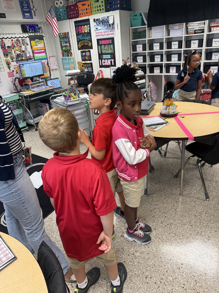 Kids in red shirts and khaki shorts stand together in a classroom, with a teacher nearby.