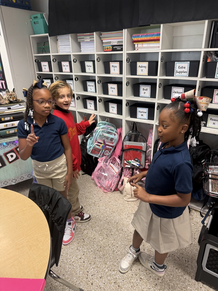 Three girls in school uniforms stand in a classroom. One holds a backpack. Shelves with storage boxes and items are behind them.