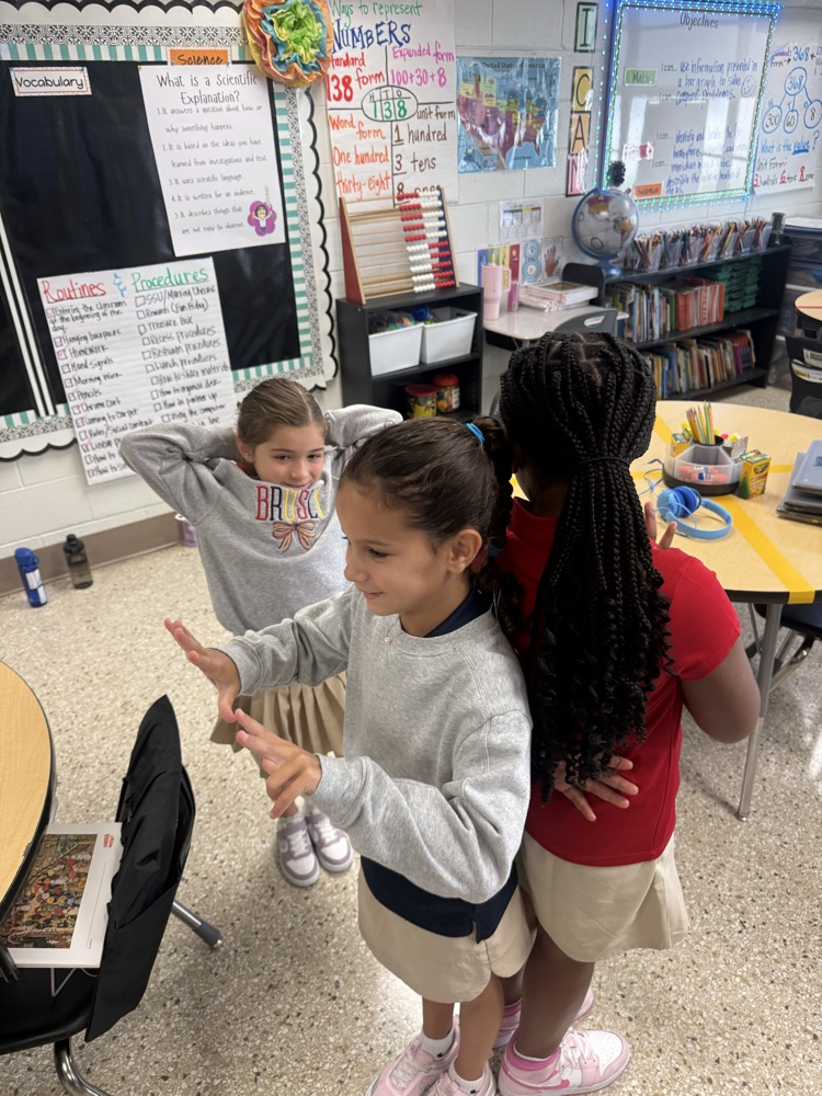 Three girls stand in a classroom with chairs and tables. One girl wears a gray sweatshirt, another in red, and the third in a blue top.