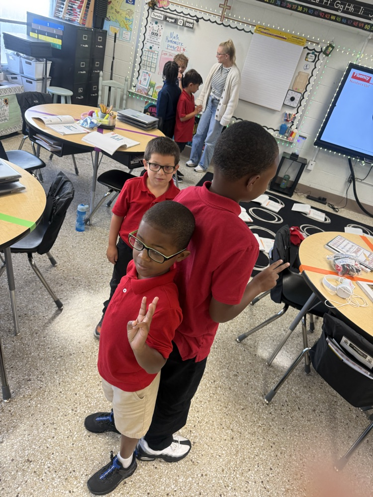 Two boys in red shirts, one with glasses, stand next to each other in a classroom.