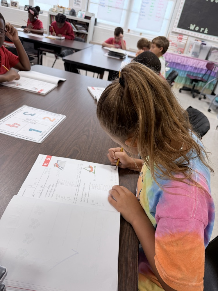 A student in a tie-dye shirt sits at a table with papers, writing on a sheet. Other students sit at tables in the background.