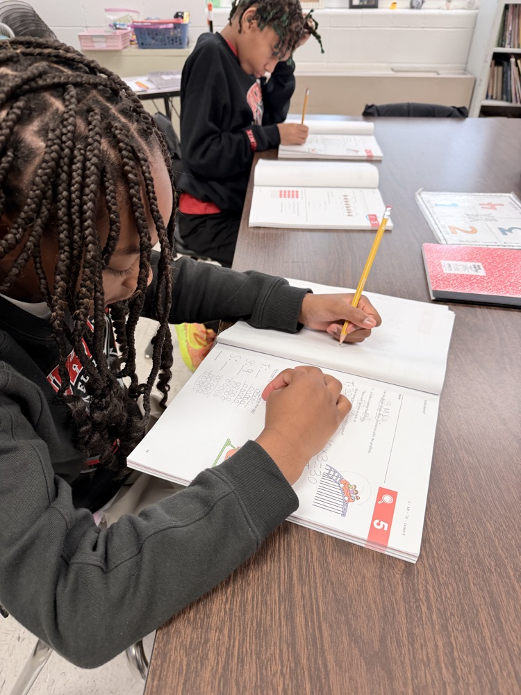 Children sit at a table writing in notebooks. One child holds a pencil and another sits behind them.
