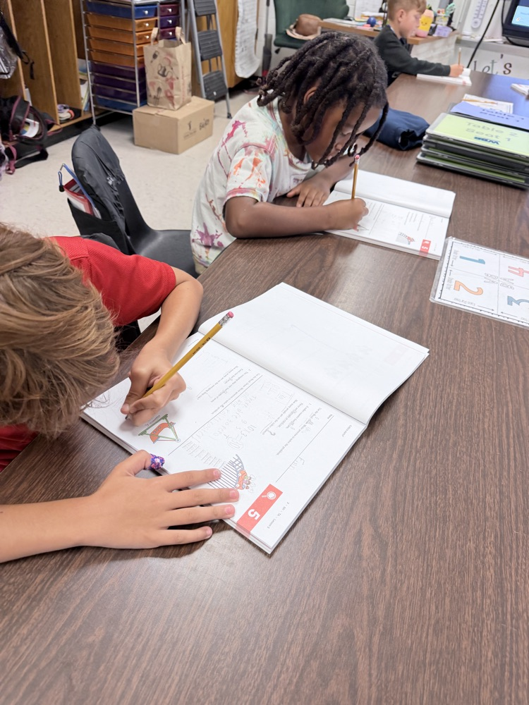 Three children work on school assignments at desks. One uses a pencil, another has a pen.