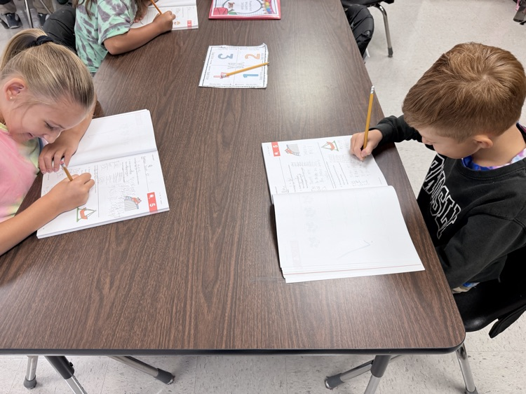 Two children at a table, one using a pencil to write in a notebook, the other using a pencil to trace lines.